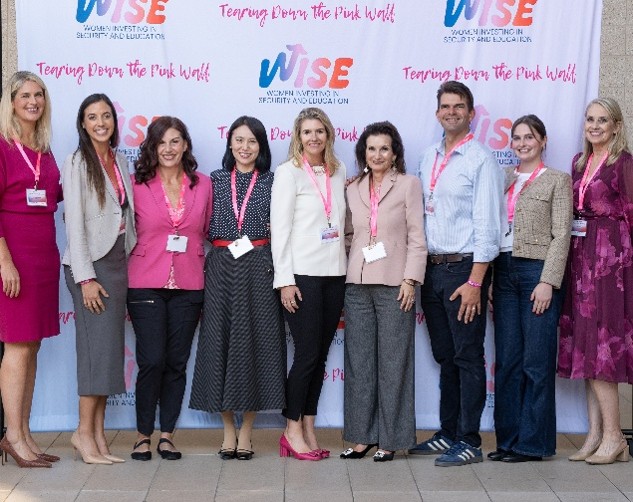 Nine conference attendees pose in front of a WISE event backdrop, wearing name badges on pink lanyards.
