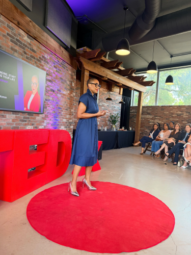 Dr. Takisha Robinson presenting on a red TEDx stage area in front of a seated audience inside a modern venue with exposed brick walls and large windows.