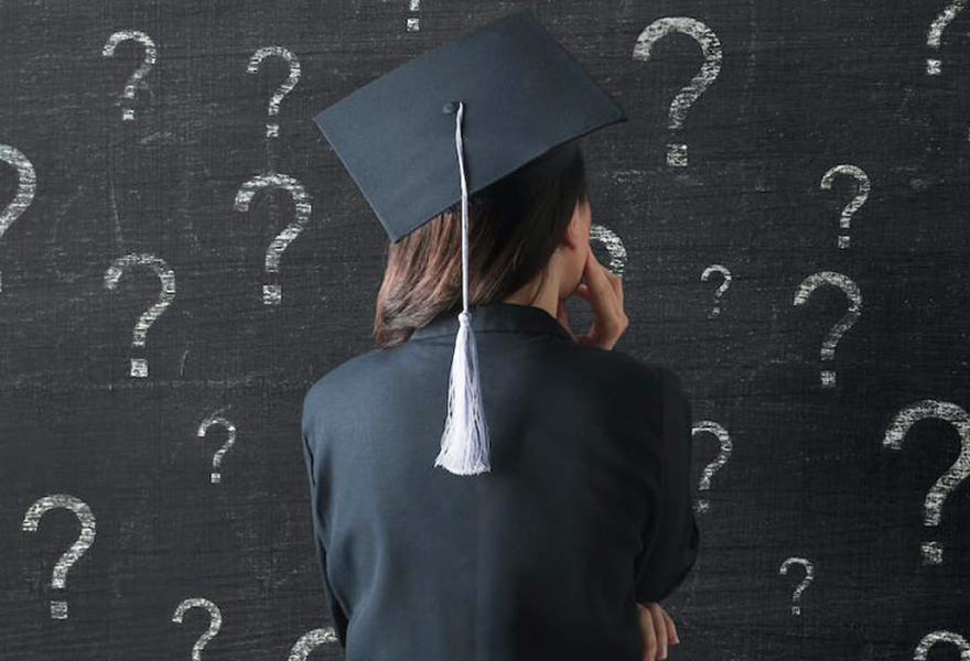 Graduating student in cap and gown looking at a chalkboard of question marks
