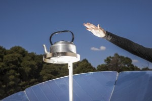 A person's hand stretches out towards a kettle which stands on a platform which is part of a large solar stove outside in the sun.